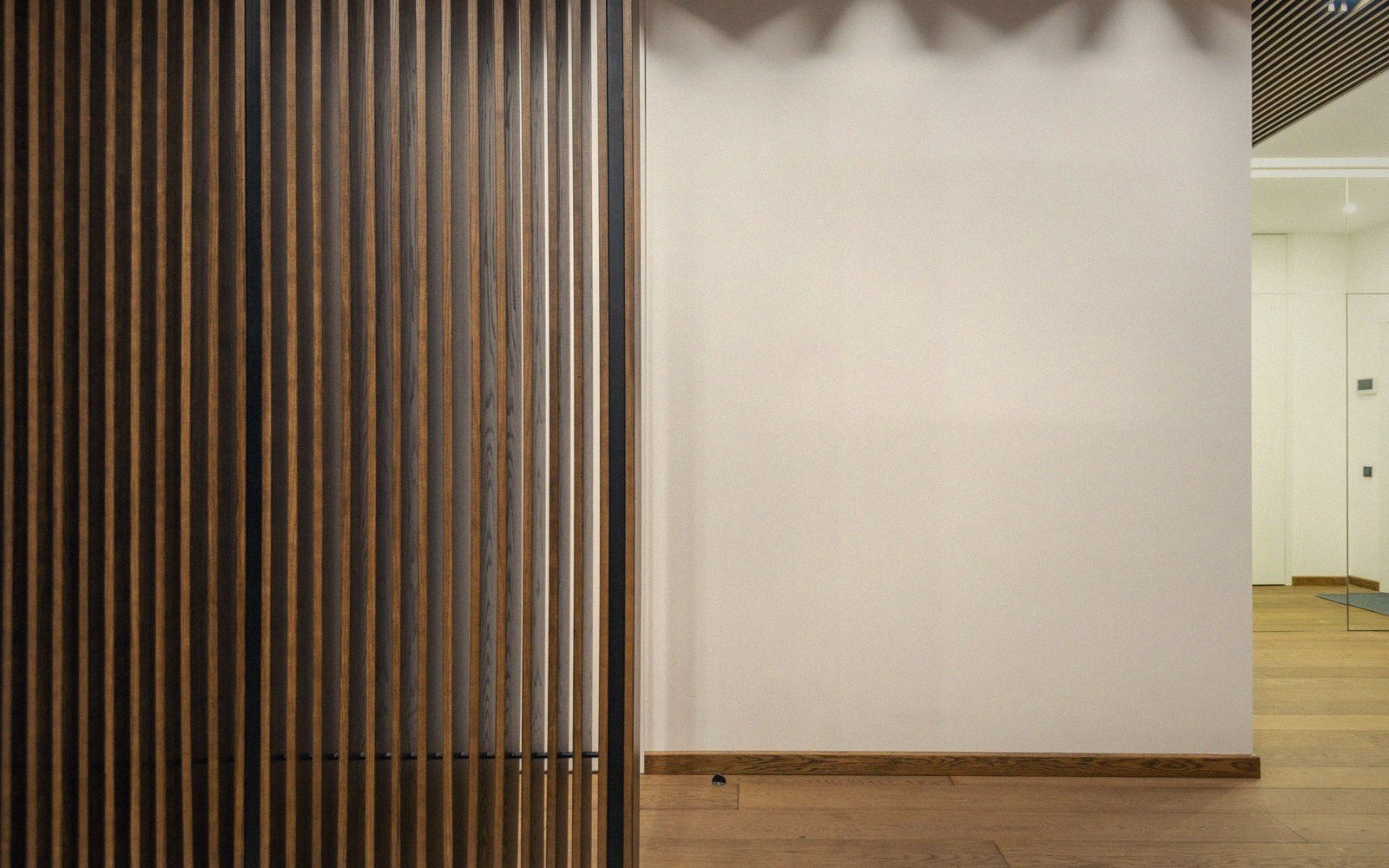 Wooden slat wall panel beside a plain white wall in a hallway with wood flooring