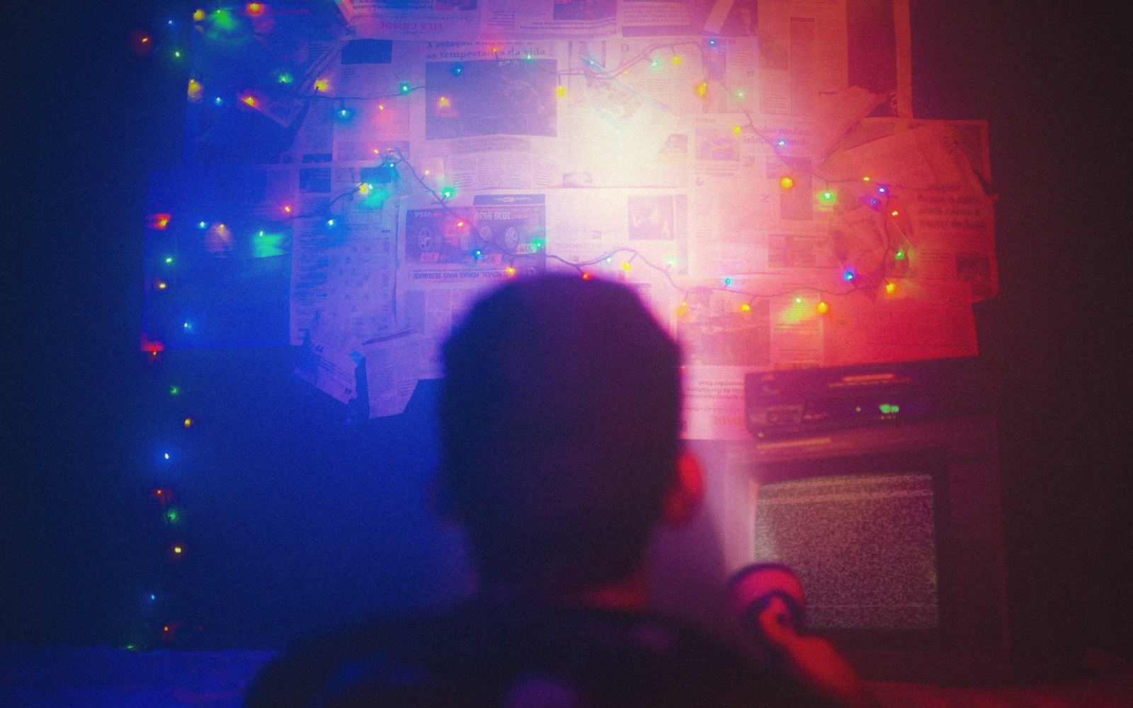 Silhouette facing a wall covered in newspaper clippings, string lights and a static-filled tv