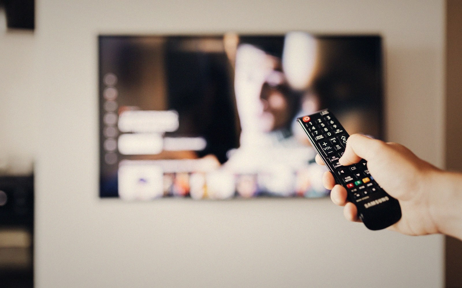 A hand points a Samsung remote control at a wall-mounted television