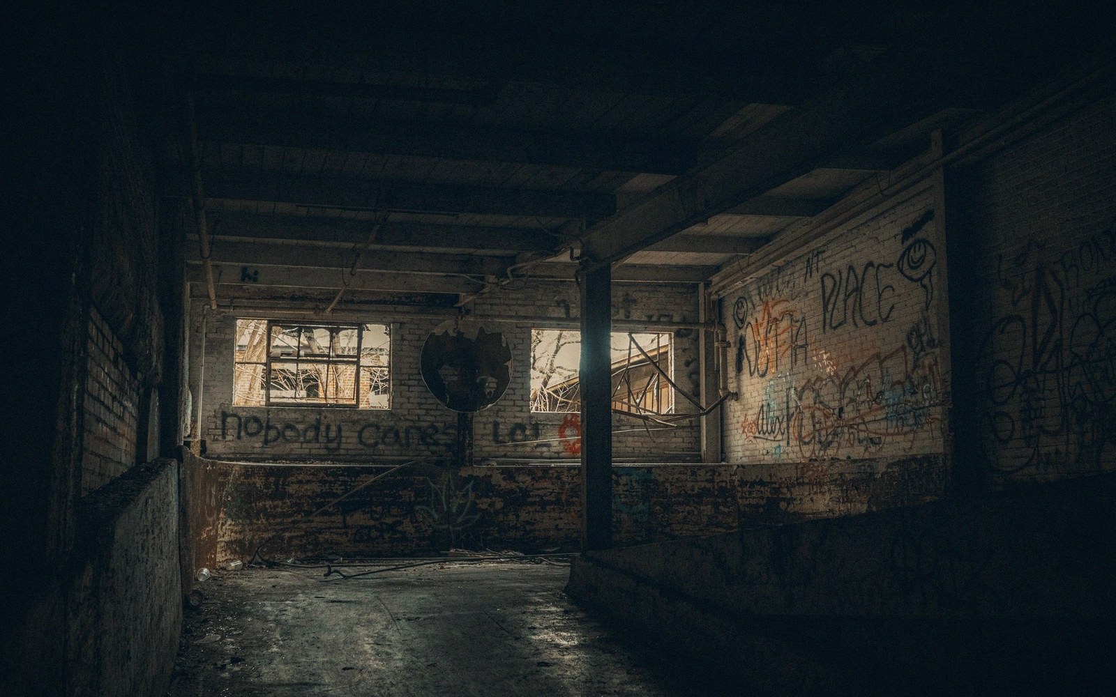 Graffiti-covered brick walls line a dim abandoned factory corridor with broken windows